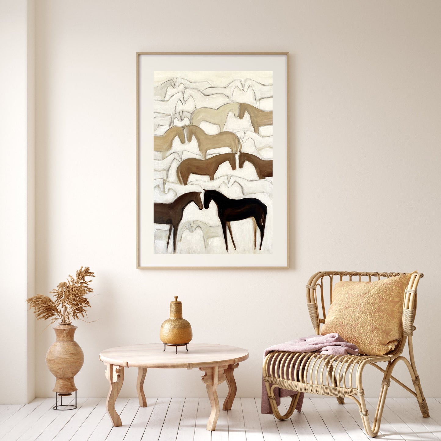 A framed artwork depicting abstract herds of horses in a mountainous landscape, displayed on a wall above a wicker chair and wooden table in a room with white walls.