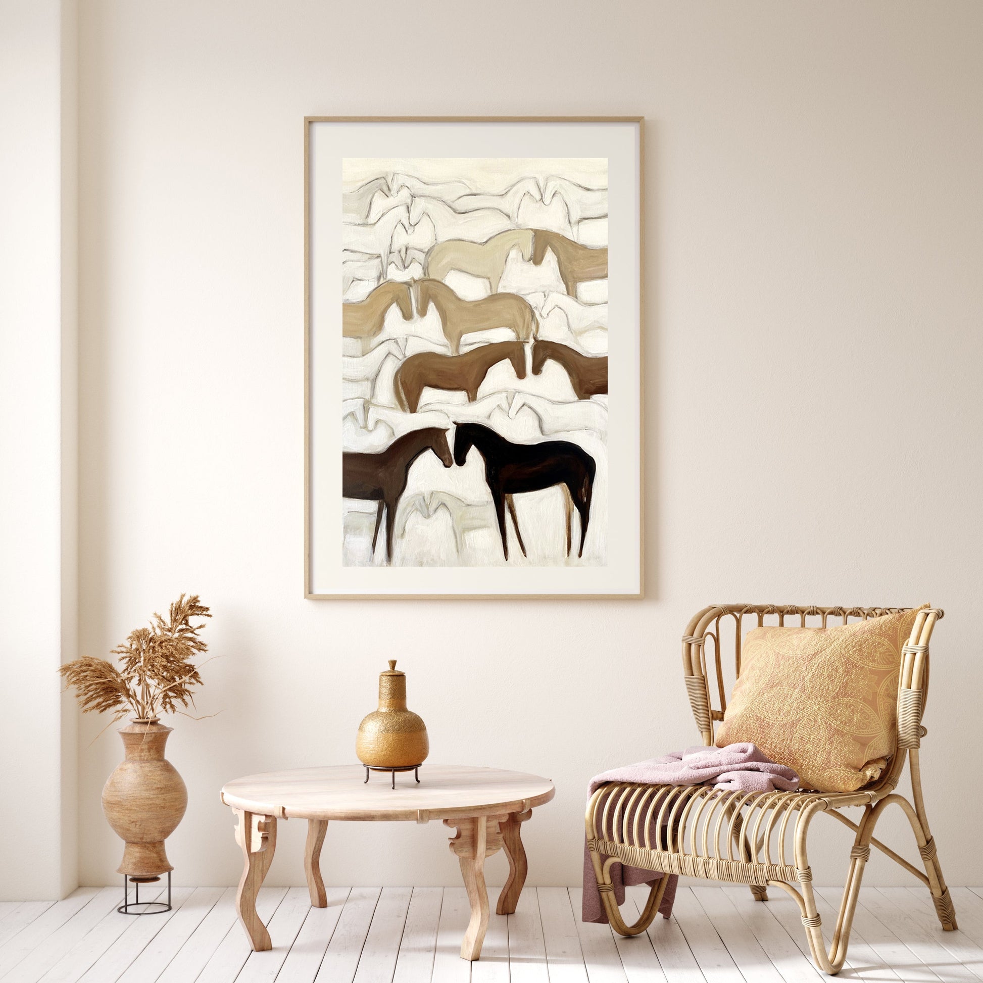 A framed artwork depicting abstract herds of horses in a mountainous landscape, displayed on a wall above a wicker chair and wooden table in a room with white walls.